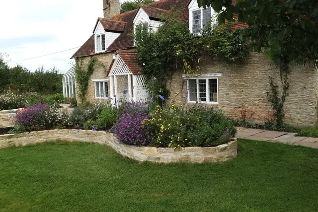 A stone cottage behind a curved, dry-stone raised border and lawn